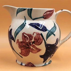 May include: A white ceramic pitcher with a floral design. The pitcher features a variety of flowers in shades of purple, pink, and green. The pitcher has a rounded body and a handle.
