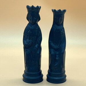 May include: A pair of dark blue chess pieces, a king and a queen, are shown. The king, on the left, holds a scepter, while the queen is on the right. Both pieces feature intricate carvings and are set against a plain background.