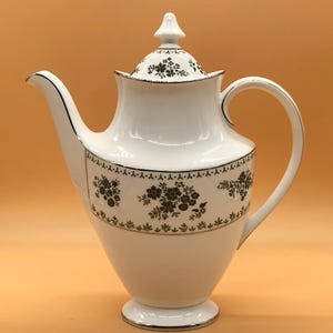 Può includere: Teiera in porcellana bianca con un motivo floreale verde e bordi dorati. La teiera ha un coperchio con un pomo in cima.
