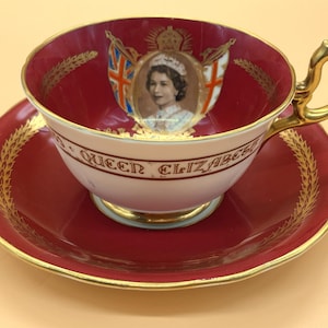 Op de afbeelding: Een vintage theekop en schotel set. De theekop en schotel zijn dieprood met gouden randen. De theekop heeft een portret van koningin Elizabeth II, de woorden "QUEEN ELIZABETH" en de Britse vlag. De handgreep is goud.
