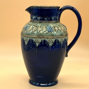 Könnte beinhalten: Ein dunkelblauer Keramikkrug mit großem Henkel und Ausguss. Der Krug hat ein dekoratives Band aus floralen und scrollartigen Verzierungen in einem helleren Blau und Grau in der Mitte. Der Krug ist etwa 25 cm hoch.