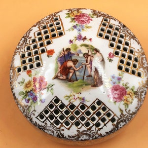 May include: A white porcelain trinket box with a gold trim and a floral design. The lid has a lattice pattern and a scene of three figures in a garden.