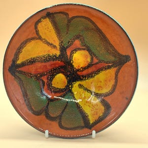 Op de afbeelding: Een decoratieve keramische plaat met een bloemmotief. De plaat heeft een verbrande oranje achtergrond met een gestileerde bloem in geel, groen en rood. De bloem is omlijnd in zwart en de rand is donkerder. Diameter: ongeveer 25 cm.