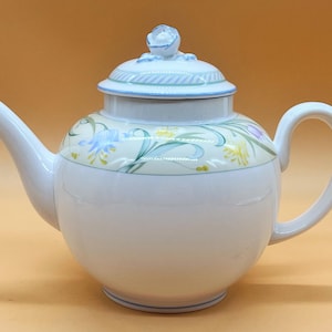 Pode incluir: Bule de chá branca com um design floral em tons de azul, amarelo e verde. A chaleira tem uma tampa com um pequeno botão floral.