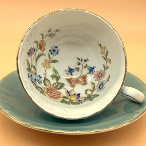 Puede incluir: Un juego de taza y platillo blancos con un diseño floral y de mariposas. La taza tiene un borde dorado y un asa blanca. El platillo es de color verde azulado claro con un borde dorado. El conjunto está sobre un fondo naranja claro.