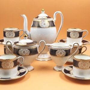 Pode incluir: Um conjunto de chá branco com detalhes dourados e pretos. O conjunto inclui uma chaleira, um jarro de creme, um açucareiro e quatro xícaras e pires.