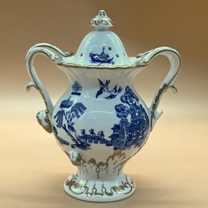 Könnte beinhalten: Eine weiß-blaue Keramikurne mit Deckel, mit einem klassischen Willow-Muster. Die Urne hat zwei Griffe und goldfarbene Akzente. Der Deckel hat einen dekorativen Knauf und kleine Löcher.