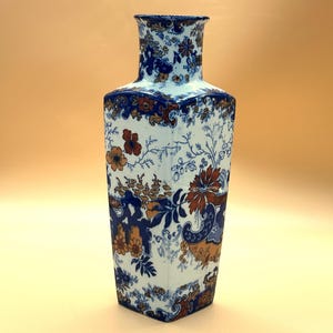 May include: A tall, rectangular ceramic vase with a flared top. The vase features a white background with a blue and orange floral pattern. The rim and base are trimmed with a dark blue border. The vase is a decorative home decor item.