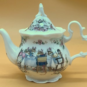 Może przedstawiać: Biały ceramiczny imbryk z pokrywką z kwiatową i mysią ilustracją. Imbryk ma zakrzywioną wylewkę i uchwyt w kształcie serca. Pokrywka jest ozdobiona napisem "Brambly Hedge".