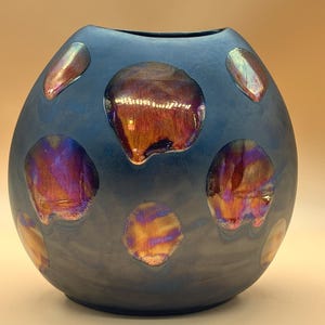 Op de afbeelding: Een afgeronde vaas met een matte donkerblauwe afwerking met verschillende onregelmatige, cirkelvormige vlekken met een glanzende, iriserende, veelkleurige metallic glans die verschuift tussen tinten koper, paars en goud over het oppervlak.