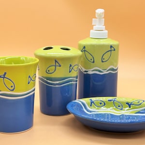 Puede incluir: Un juego de baño con un esquema de color verde y azul. El juego incluye un portacepillos de dientes, un dispensador de jabón, un vaso y un jabonera. Todas las piezas presentan un diseño de olas con pequeños peces.