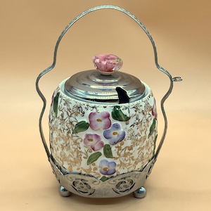 Op de afbeelding: Een vintage suikerpot met een bloemmotief. De keramische kom heeft roze en paarse bloemen met groene bladeren op een crèmekleurige achtergrond. Het heeft een zilveren deksel en handvat en staat op een zilveren voet met drie poten. Een kleine lepel rust erin.