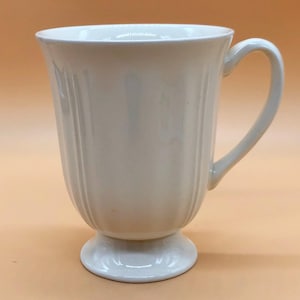 Può includere: Una tazza in ceramica bianca con base a piedistallo e design scanalato. La tazza ha un manico ed è perfetta per servire caffè o tè.