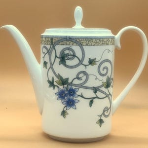 May include: White ceramic teapot with a curved spout and handle. The teapot features a decorative design of blue flowers, green leaves, and gray swirls. A matching lid with a small knob completes the design. The teapot is likely for serving tea.