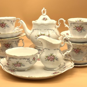 Puede incluir: Un juego de té de porcelana blanca con un diseño floral. El juego incluye una tetera, una jarra de crema, un azucarero, cuatro tazas y cuatro platillos. El juego está decorado con flores rosas y azules y tiene un borde dorado.