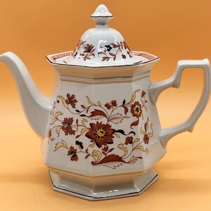 May include: White teapot with a floral design in shades of brown and yellow. The teapot has a lid with a knob on top and a handle on the side.