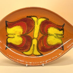 Puede incluir: Un plato de cerámica ovalado para servir, de color naranja cálido. Presenta un diseño central simétrico y abstracto en amarillo, rojo y marrón. El diseño es de estilo moderno de mediados de siglo.