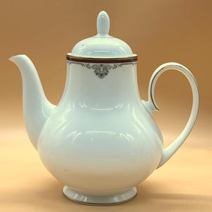 Pode incluir: Bule de porcelana branco com tampa e alça curva. O bule tem um bico e uma faixa decorativa de acabamento vermelho escuro e dourado ao redor da tampa. A alça e os acabamentos têm detalhes dourados.
