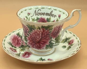 May include: A white teacup and saucer set with a floral design featuring large pink chrysanthemums and green leaves. The teacup has the word "November" written on the rim. The set has gold trim.