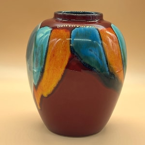 Könnte beinhalten: Eine bauchige Keramikvase mit einer glänzenden, tiefroten Grundfarbe und markanten vertikalen Pinselstrichmustern in leuchtendem Orange und hellem Türkisblau. Die Vase hat eine schmale, kreisförmige Öffnung oben und eine glatte, reflektierende Oberfläche.