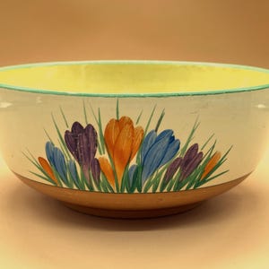 May include: A round ceramic bowl with a pale yellow interior and a white exterior featuring a hand-painted floral design of purple, orange, and blue crocus flowers with green stems. The base of the bowl is unglazed terracotta brown.