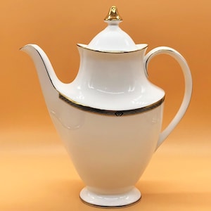 May include: White porcelain coffee pot with a gold lid and a black and gold trim around the top. The pot has a rounded body and a spout on the side.