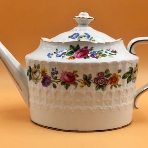 Może przedstawiać: Biały porcelanowy czajnik z kwiatowym wzorem i złotą obwódką. Czajnik ma pokrywkę z gałką i uchwytem.