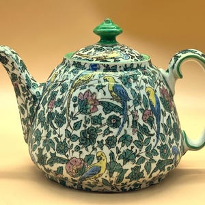 May include: A ceramic teapot with a green handle, lid, and spout. The teapot is decorated with a pattern of colorful birds, flowers, and green foliage on a cream background. The teapot has a rounded shape.