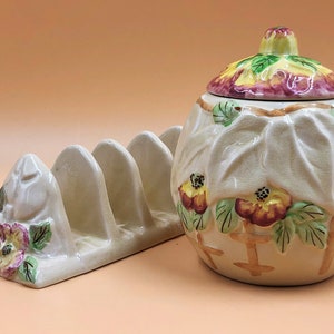 Puede incluir: Un mantequillero de cerámica vintage con tapa, con un diseño floral con flores amarillas, rosas y verdes. El mantequillero es blanco con un glaseado crema y tiene un tostadora de cuatro ranuras.