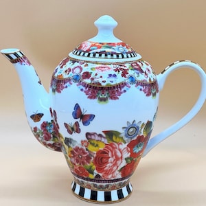 Pode incluir: Bule de porcelana branco com um design floral e de borboletas. O bule apresenta uma alça e um bico curvos, uma tampa com um botão e uma base listrada em preto e branco. O padrão floral inclui rosas vermelhas, borboletas azuis e outras flores coloridas.