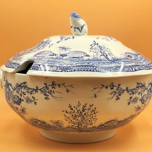 May include: A white and blue ceramic soup tureen with a lid and ladle slot. The tureen features a blue landscape design with trees and animals. It has two handles and a decorative finial on the lid.