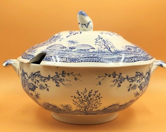 Op de afbeelding: Een wit en blauw keramieken soepterrine met deksel en sleuf voor een lepel. De terrine heeft een blauw landschapsontwerp met bomen en dieren. Het heeft twee handvatten en een decoratieve knop op het deksel.