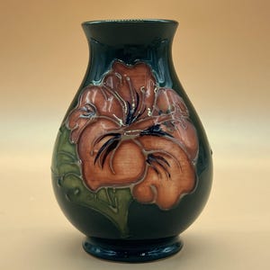 Puede incluir: Un jarrón de cerámica verde oscuro con cuerpo bulboso y borde acampanado. El frente presenta un diseño floral en relieve en tonos terracota y verde apagado, con contornos oscuros que definen los pétalos y las hojas sobre el acabado brillante.