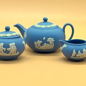 Puede incluir: Un juego de té de porcelana azul y blanco con tetera, jarra de crema y azucarero. Cada pieza presenta un diseño en relieve blanco de niños jugando en un jardín.