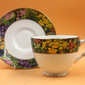 Op de afbeelding: Een wit theekopje en schotel met een bloemenpatroon met gele narcissen, paarse hyacinten en andere wilde bloemen. De rand van de schotel en het kopje zijn afgewerkt met goud.