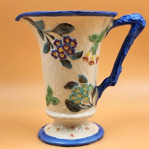 May include: A white ceramic pitcher with a blue and white floral design and a blue wooden handle. The pitcher has a pedestal base with a blue rim.