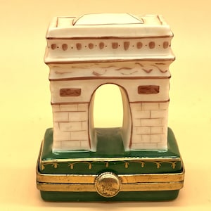 Op de afbeelding: Een witte porseleinen sieradenkistje in de vorm van de Arc de Triomphe in Parijs. De kist heeft een groene basis met gouden versieringen en een scharnierend deksel.