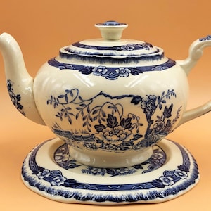 May include: A white teapot with a blue and white floral design and a matching saucer. The teapot has a round body and a spout on the side. The saucer has a scalloped edge.