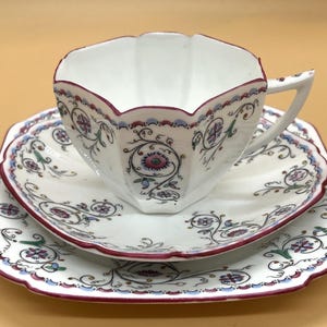 Könnte beinhalten: Weißes Tee-Set mit Blumendesign. Die Tasse hat eine einzigartige Form mit einem gewellten Rand, und die Untertasse ist quadratisch mit passendem Muster. Das Set hat rote, blaue und violette Akzente.