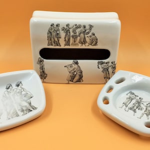 Puede incluir: Un juego de baño de cerámica blanca con un porta pañuelos, un jabonera y un porta cepillos de dientes. Cada pieza presenta un diseño en blanco y negro de figuras en procesión.