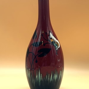 Peut inclure: Un vase en céramique rouge foncé, haut et élancé, doté d'un col étroit. La base présente des brins d'herbe peints en vert foncé et vert clair. Le corps arbore des silhouettes noires de plantes aquatiques et un motif floral blanc, jaune et vert.