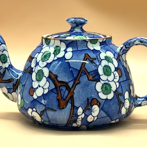 Puede incluir: Tetera de cerámica azul con tapa, decorada con motivos florales blancos y verdes y ramas marrones. La tetera tiene un asa curva y un pico con ribete dorado.