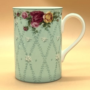Può includere: Una tazza alta in ceramica azzurra con manico bianco. La tazza è decorata con un motivo floreale di rose nei toni del rosa, giallo e bordeaux. Il design include un delicato motivo a vite e fiori bianchi.