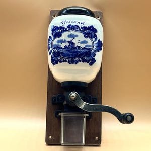 Könnte beinhalten: Eine Wandkaffeemühle mit einem weißen Keramikbehälter, der mit blauen Blumenmustern und einer Windmühlenillustration verziert ist und die Aufschrift Holland trägt. Sie umfasst eine dunkle Holzrückwand, eine Metallkurbel und einen Auffangbehälter.