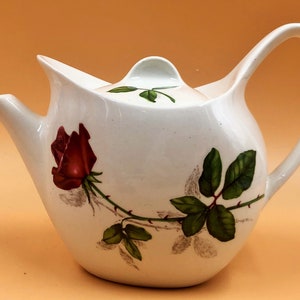May include: White ceramic teapot with a red rose and green leaves design. The teapot has a curved handle and a spout.