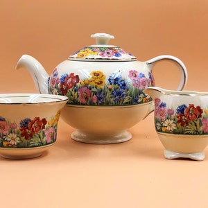 Pode incluir: Um conjunto de chá floral vintage com bule, jarra de creme e açucareiro. O conjunto apresenta um fundo branco com um padrão floral colorido.