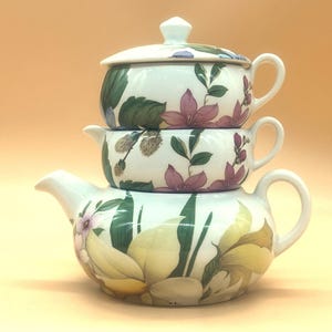 May include: Stacked white ceramic teaware set with floral designs. The set includes a teapot, a covered bowl, and a small covered dish. The floral pattern features pink, yellow, and green accents.