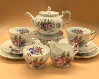 May include: A vintage tea set featuring a teapot, two teacups with saucers, a creamer, and a sugar bowl. The white porcelain set is adorned with floral patterns in pink, purple, yellow, and blue, with gold trim. A classic tea party set.