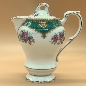 Puede incluir: Tetera de porcelana blanca con tapa, con una banda decorativa verde y dorada. La tetera está adornada con motivos florales en tonos rosa, morado y amarillo. El asa y el borde tienen adornos dorados.