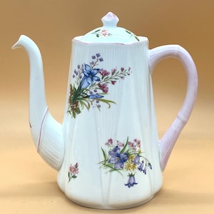 May include: White ceramic coffee pot with a floral design featuring blue, purple, and pink flowers. The handle and accents are light pink. The lid has a small pink finial and floral details. The pot has a tall, slender spout.